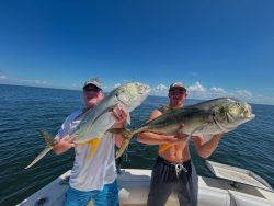 Two people on a boat each hold a large fish, with open water and a clear sky in the background.