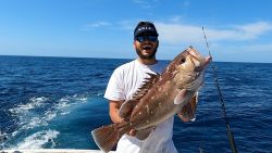 Man wearing sunglasses and a visor holds a large fish on a boat in the ocean under a clear blue sky.