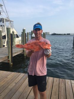 Man standing on a dock holding a large reddish fish with both hands; boats and water visible in the background.
