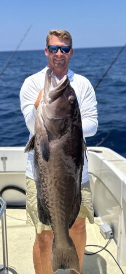 Man on a boat holding a large fish with the ocean and fishing rods in the background.