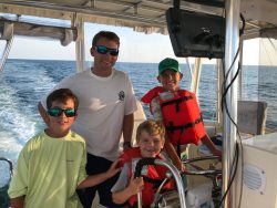 Four people, including three boys in life jackets, pose on a boat with open water in the background on a sunny day.
