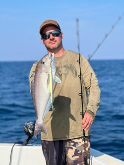 Man standing on a boat holding a fishing rod and displaying a freshly caught fish with the ocean in the background.