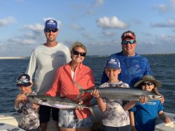 Five people on a boat smiling and holding two large fish, with ocean and sky in the background.