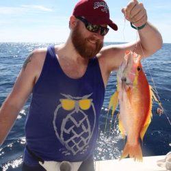 Man in sunglasses and a red cap holding up a colorful fish on a boat with the ocean in the background.