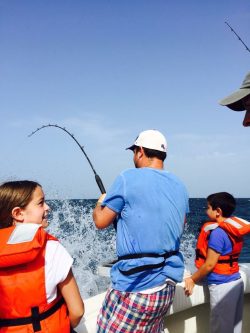 Man in blue shirt fishing from a boat with two children in orange life jackets and water splashing nearby.