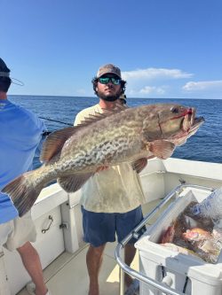 Man holding a large fish on a boat, with ocean and blue sky in the background. Cooler with fish visible in foreground.