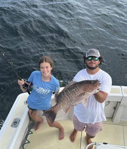 Two people on a boat; one holds a fishing rod while the other displays a large fish caught from the ocean.