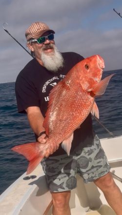 Man standing on a boat holds a large red snapper fish with the ocean and fishing rods in the background.