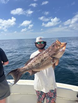 Man on a boat holds a large fish with the ocean and cloudy blue sky in the background.
