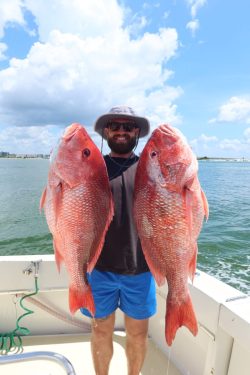 Man on a boat holding two large red fish, wearing a hat, sunglasses, and blue shorts with water and sky in the background.