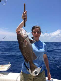 A woman on a boat holds up a large fish she caught, with blue ocean and sky in the background.