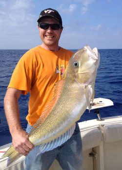 Man in an orange shirt holding a large fish on a boat with the ocean in the background.