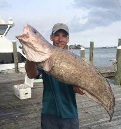 Man standing on a dock holding a large fish with boats and water in the background.