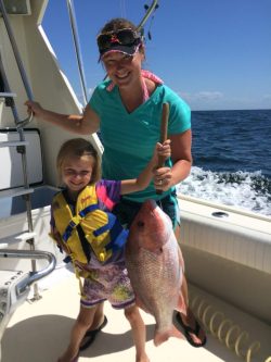 An adult and child on a boat hold a large fish together, both smiling with the ocean in the background.