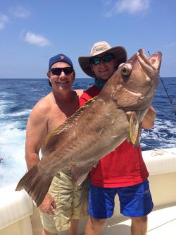 Two people on a boat holding a large fish with the ocean and blue sky in the background.