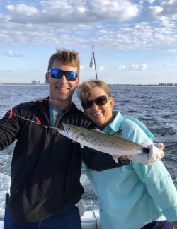 Two people on a boat smiling and holding a freshly caught fish with the ocean and a partly cloudy sky in the background.