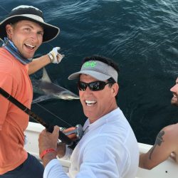 Three men on a boat catching a shark, smiling at the camera while holding a fishing rod over the water.