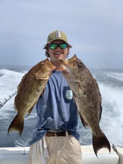 Person on a boat holds up two large fish, standing in front of the ocean with waves in the background.