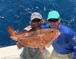 Two men on a boat holding a large reddish-brown fish with the ocean in the background.