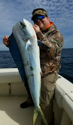 Person in camo gear holding a large dolphinfish (mahi-mahi) on a boat with the ocean and sky in the background.