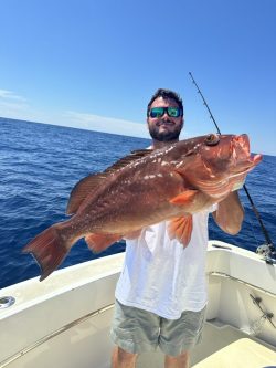 Man on a boat holding a large grouper fish with the ocean and a fishing rod in the background.