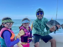 Two girls and a man on a boat smile at the camera; the man is holding a fish. The ocean is in the background.
