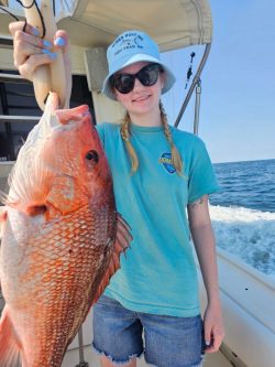 Person holding a large red fish on a boat, wearing a blue shirt, sunglasses, and a hat; ocean in the background.