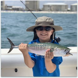 Child wearing a hat and sunglasses holding a large fish on a boat with buildings and water in the background.