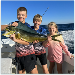 Three children on a boat hold a large fish together, smiling, with the ocean and blue sky in the background.