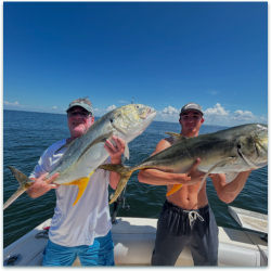 Two men standing on a boat hold large fish, with open water and blue sky in the background.