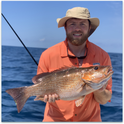 A man in an orange shirt and hat holds a large fish on a boat with the ocean in the background.