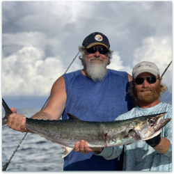 Two men on a boat hold a large, freshly caught fish with the ocean and cloudy sky in the background.